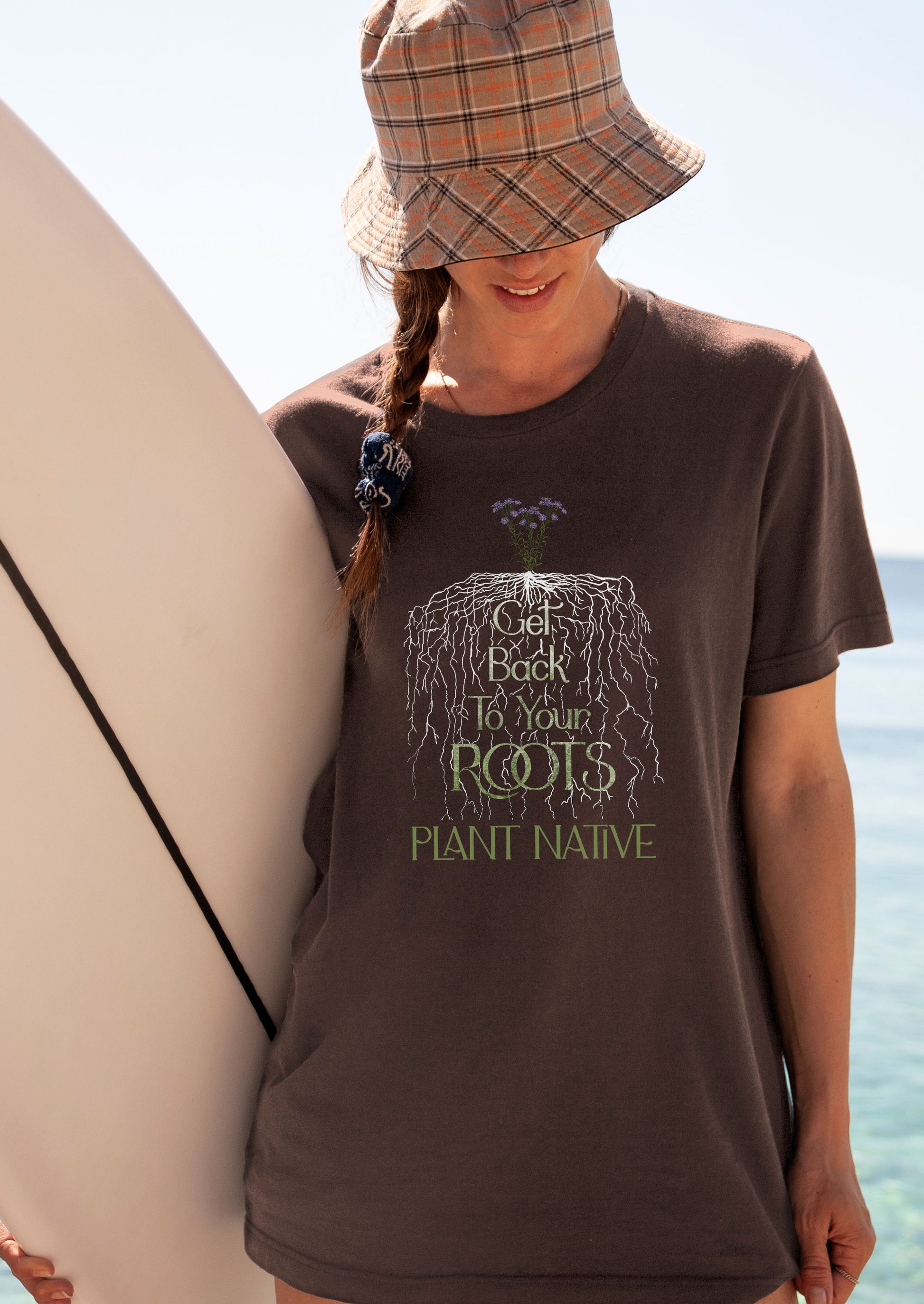 Girl holding surf board wearing a brown shirt with roots from a native plant that says, Get Back to Your Roots. Plant native"